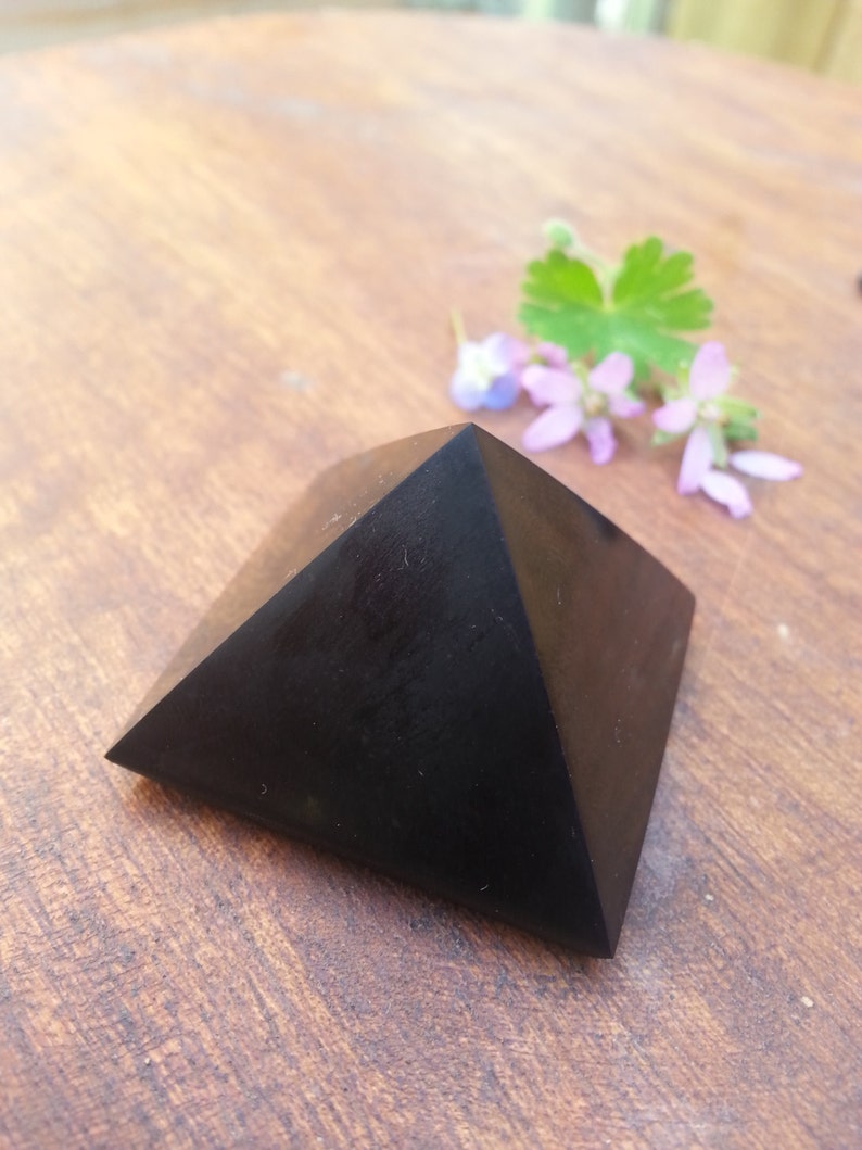May include: Black obsidian pyramid crystal on a wooden surface. The pyramid is smooth and polished.