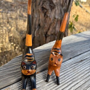 May include: Two wooden cat figurines with elongated bodies and tails. One cat is black with orange stripes, the other is orange with black stripes. Both have detailed facial features and stand on four legs.