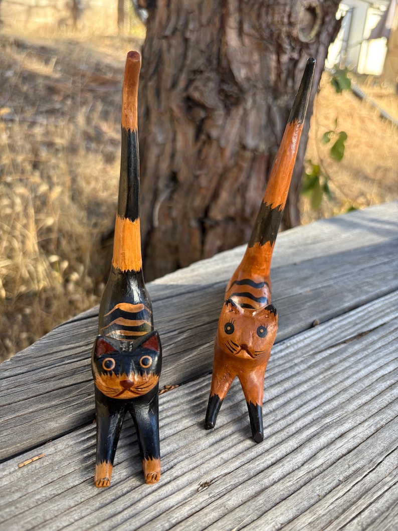 May include: Two wooden cat figurines with elongated bodies and tails. One cat is black with orange stripes, the other is orange with black stripes. Both have detailed facial features and stand on four legs.