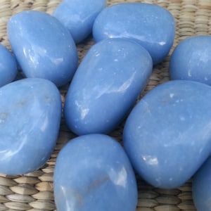 May include: A collection of smooth, oval-shaped blue stones. The stones have a glossy finish and vary slightly in size. They are arranged on a woven, light brown surface. The stones are a soft, light blue color.