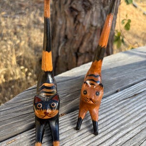 May include: Two wooden cat figurines with upright tails. One cat is black with orange stripes, the other is orange with black stripes. Both have detailed facial features and stand on four legs. The cats are hand-carved and painted.
