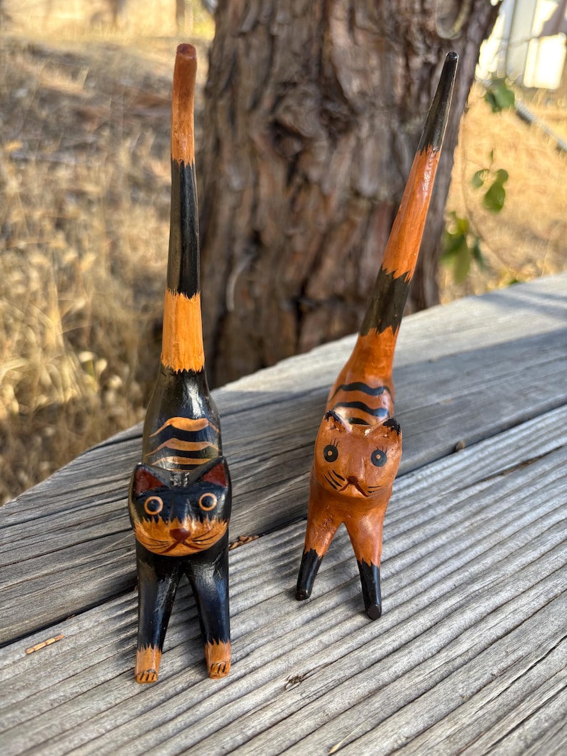 May include: Two wooden cat figurines with upright tails. One cat is black with orange stripes, the other is orange with black stripes. Both have detailed facial features and stand on four legs. The cats are hand-carved and painted.