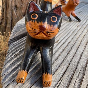 May include: Hand-carved wooden cat figurine with a black body and orange and black face. The cat has large, round eyes and a detailed, painted design. Another cat figurine is visible in the background. The figurines are on a wooden surface.