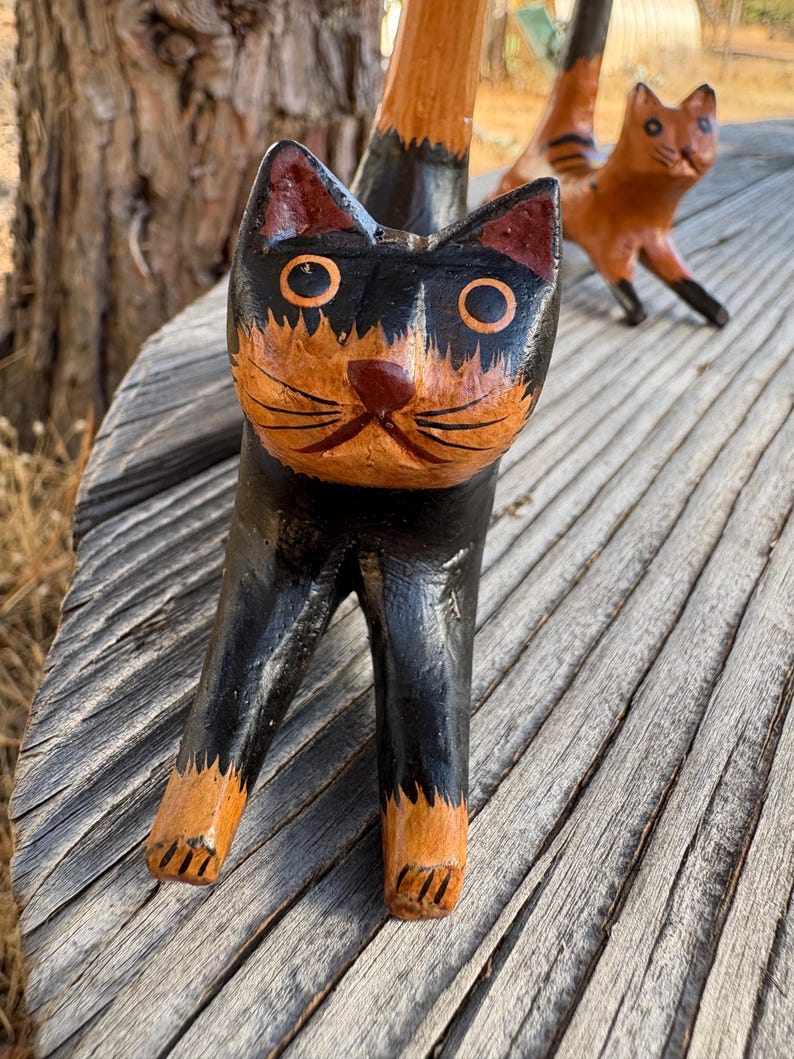 May include: Hand-carved wooden cat figurine with a black body and orange and black face. The cat has large, round eyes and a detailed, painted design. Another cat figurine is visible in the background. The figurines are on a wooden surface.