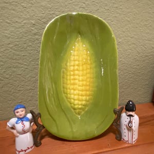 May include: A green ceramic dish shaped like an ear of corn. The dish has a yellow corn cob design in the center. The dish is on a wooden surface with two small ceramic figurines on either side.