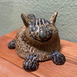 May include: A whimsical ceramic sculpture of a creature with a smiling face, large ears, and textured fur. The sculpture is primarily brown with darker accents on the face and paws. It sits on a wooden surface.