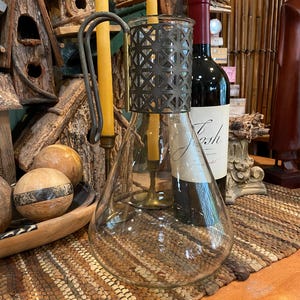 May include: A clear glass bottle with a metal cage around the top. The bottle is sitting on a woven rug with a wooden surface underneath. There are two yellow candles and a bottle of wine in the background.