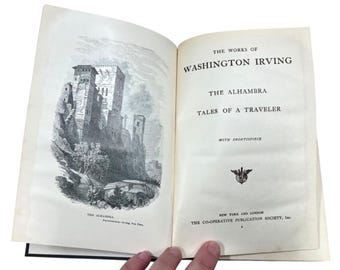 May include: Open book with the title "The Works of Washington Irving" and "The Alhambra" visible. The left page features a detailed black and white illustration of a castle. The book is held open by a person.