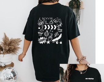 Op de afbeelding: Zwart T-shirt met witte krabbels en tekst, waaronder "Things I can't" en "DCXVII". De achterkant van het shirt heeft verschillende krabbels, waaronder een veer, een ster, een vredesteken en een klok. De voorkant van het shirt heeft de tekst "One Direction" in wit.