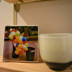 May include: A colorful photo of a bunch of balloons tied to a lamppost. The photo is framed and sitting on a wooden shelf with a white candle in a clear glass jar.