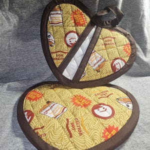 Peut inclure: Deux maniques en forme de cœur brunes avec un fond jaune et un motif de nouilles ramen, un visage de dessin animé et le texte "Rich Flavor" et "Caution Hot".