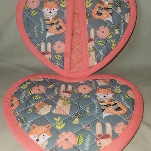 May include: A heart-shaped quilted pot holder with a gray background featuring a repeating pattern of foxes, rabbits, hedgehogs, mushrooms, and flowers in orange, green, and white. The edges are finished with a solid coral-colored fabric trim.