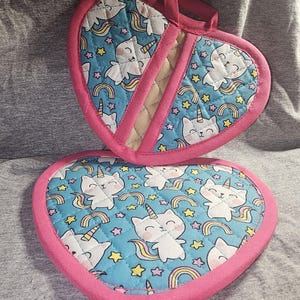 Puede incluir: Dos agarraderas para ollas en forma de corazón con un borde rosa y un fondo azul. Las agarraderas presentan un patrón repetitivo de gatos blancos con cuernos de unicornio y colas de arcoíris. Los gatos están rodeados de estrellas amarillas y arcoíris.
