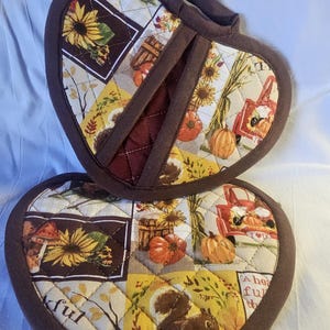 May include: A pair of brown heart-shaped pot holders with a quilted design. The pot holders feature a patchwork of autumn-themed fabric with images of sunflowers, pumpkins, squirrels, and a red truck. The text "Thankful" is visible on one of the pot holders.