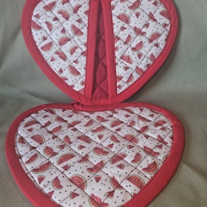Peut inclure: Une paire de maniques matelassées en forme de cœur avec un tissu blanc à motif répétitif de tranches de pastèque et de pépins noirs. Les bords sont finis avec un biais en tissu rouge uni, et la pièce supérieure inclut une petite boucle rouge.