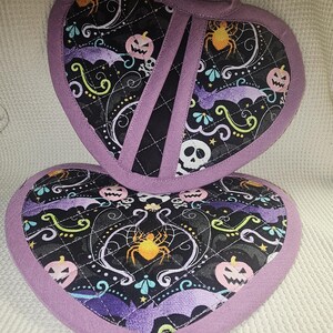 Könnte beinhalten: Zwei herzförmige Topflappen in Lila und Schwarz mit Halloween-Motiv. Der Stoff hat einen schwarzen Hintergrund mit Fledermäusen, Kürbissen, Spinnen und Totenköpfen in verschiedenen Farben.