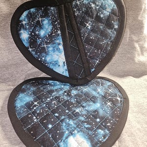 May include: A pair of heart-shaped pot holders with a galaxy print. The pot holders are quilted and have a black border.