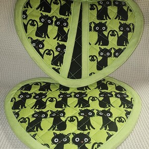 Peut inclure: Deux maniques en forme de cœur vertes avec des motifs de chats noirs. Les maniques ont une bordure en tissu noir et une poignée en tissu noir.