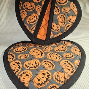 May include: Two black and orange heart-shaped pot holders with a pattern of orange jack-o'-lantern faces on a gray background.