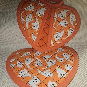 Pode incluir: Um conjunto de dois pegadores de panela acolchoados laranja e branco com um padrão de fantasma. Os pegadores de panela são em forma de coração e têm um laço para pendurar.