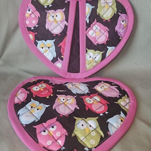 May include: A heart-shaped quilted potholder with a black background featuring a colorful owl pattern in shades of pink, green, blue, and yellow. The edges are finished with a solid pink fabric trim, and it includes a small fabric hanging loop.