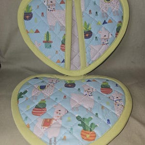 Peut inclure: Un dessous-de-plat matelassé en forme de cœur avec un tissu bleu clair orné d'un motif répétitif de lamas blancs, de cactus en pot colorés et de petits triangles. Les bords sont finis avec un biais vert clair et une boucle de suspension.