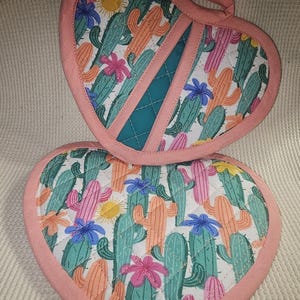 Peut inclure: Deux maniques en forme de cœur roses avec un motif de cactus et de fleurs. Les maniques sont matelassées et la manique du dessus a un accent en tissu turquoise.