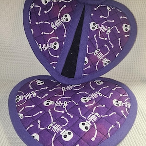 Peut inclure: Une paire de maniques en forme de cœur de couleur violette avec un motif de squelette blanc. Les maniques sont matelassées et ont une doublure en tissu noir.
