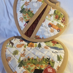 May include: Two heart-shaped quilted pot holders with a fall theme. The pot holders feature a brown quilted border, a white background, and a colorful fall scene with pumpkins, trees, and a red barn. One pot holder has the text "hello fall" and the other has the text "to fall".