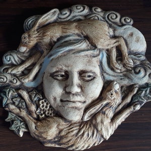 May include: A ceramic wall sculpture depicting a woman's face surrounded by three hares. The sculpture is painted in shades of brown, white, and blue, with a textured surface.