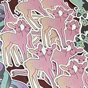 May include: A collection of stickers featuring various designs, including pastel-colored snakes, deer with two heads, and green and black cats. The stickers are all on a brown background.