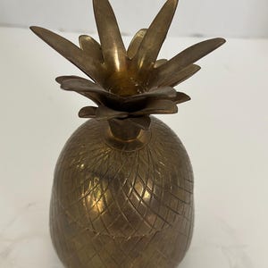 May include: A brass pineapple-shaped container with a removable lid. The body features a textured, cross-hatched diamond pattern, while the top lid is shaped like pointed pineapple leaves. The metal has a warm, aged, golden-brown patina finish.