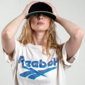 Op de afbeelding: Zwarte baseballcap met een groene klep wordt boven het hoofd gehouden. De persoon draagt een wit t-shirt met het blauwe Reebok-logo. Het shirt heeft korte mouwen.