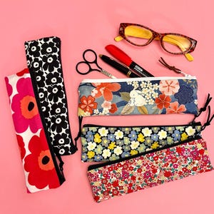May include: Various pencil cases with floral patterns in red, black, and blue. The image also shows eyeglasses, scissors, and pens, all arranged on a pink surface.