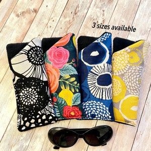 May include: Four different patterned fabric eyeglass cases with black trim. The cases are lined up in a row on a wooden surface. The text "3 sizes available" is written in white on a black background above the cases. A pair of black sunglasses is in the foreground.
