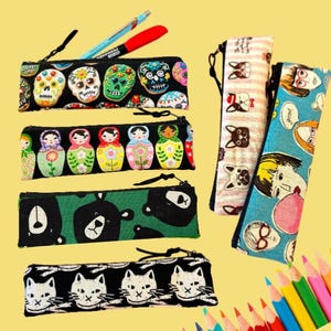 May include: Collection of pencil cases with various patterns, including sugar skulls, nesting dolls, bears, cats, and dog faces. The cases are black with colorful designs and have black zippers. Two pens are visible on top of one case.