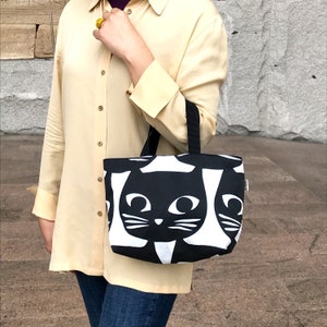 Puede incluir: Una bolsa de mano de color blanco y negro con un patr&oacute;n repetido de caras de gatos negros. La bolsa tiene dos asas negras.