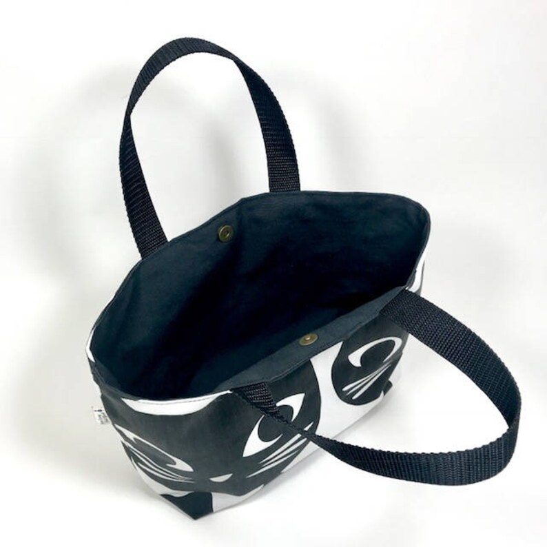 Puede incluir: Bolso de mano blanco y negro con un dise&ntilde;o de cara de gato. El bolso tiene asas negras y un forro negro.