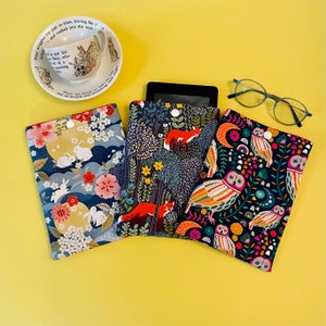 May include: Three fabric e-reader cases with button closures, featuring colorful patterns. One case has a floral and rabbit design, another has foxes in a forest, and the third has owls. A teacup and glasses are also visible.