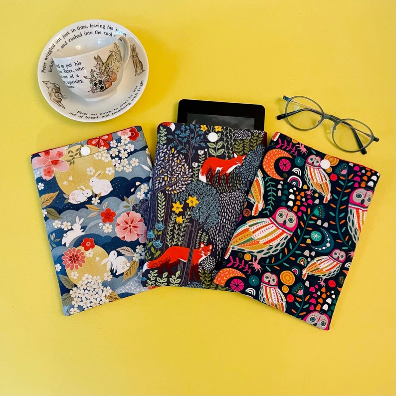 May include: Three fabric e-reader cases with button closures, featuring colorful patterns. One case has a floral and rabbit design, another has foxes in a forest, and the third has owls. A teacup and glasses are also visible.