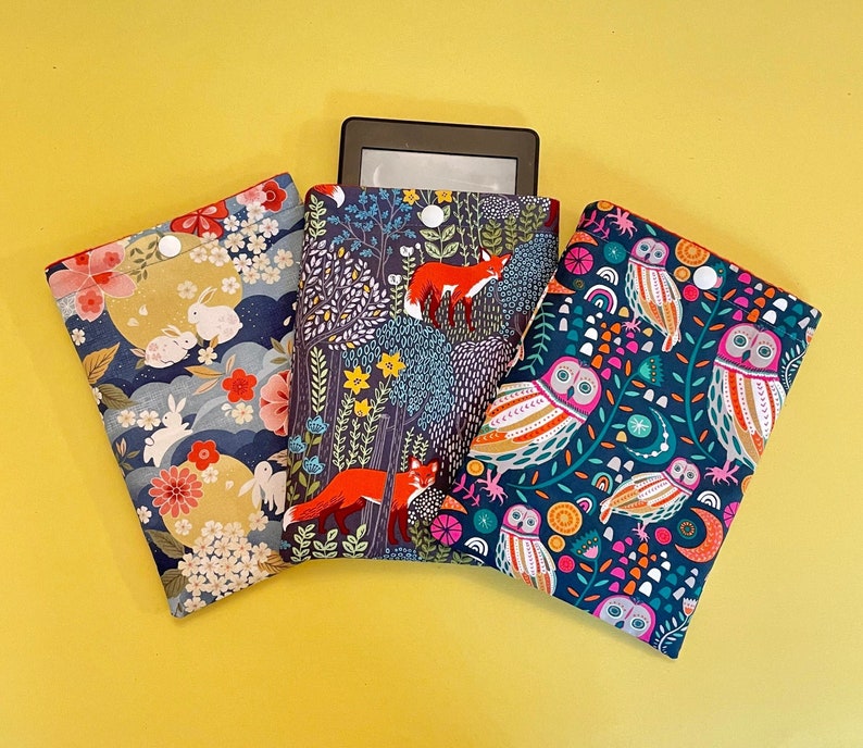May include: Three fabric e-reader sleeves with colorful patterns. One features rabbits and flowers, another has foxes in a forest, and the third showcases owls. Each sleeve is designed to protect e-readers.