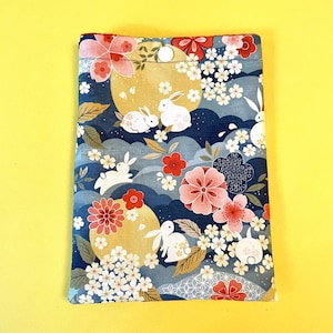May include: A rectangular fabric case with a Japanese-inspired design. The pattern features white rabbits, full moons, cherry blossoms, and stylized clouds in shades of blue, red, and gold. The case is secured with a white button.