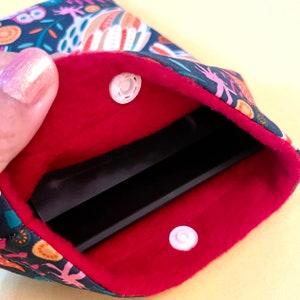 May include: A small, open pouch with a red interior and a patterned exterior featuring colorful floral and abstract designs. The pouch is secured with two white snap closures. The pouch is held open, revealing a black rectangular object inside.