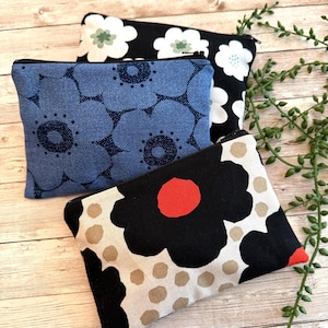 May include: Three rectangular zippered pouches made of fabric. One pouch features a blue floral pattern, another has a black background with white flowers, and the third displays a black flower with a red center on a cream background with tan dots.