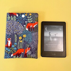 May include: A Kindle e-reader next to a fabric book cover. The cover features a dark blue background with a repeating pattern of orange foxes, trees, flowers, and a moon. The Kindle screen displays text that reads, "Your next great read is waiting."