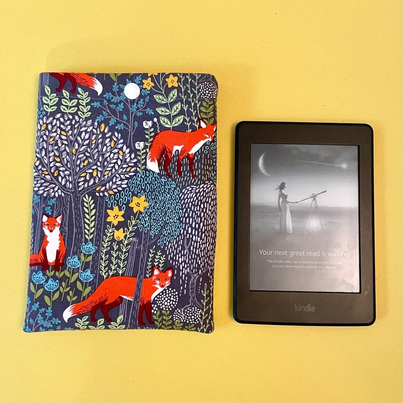 May include: A Kindle e-reader next to a fabric book cover. The cover features a dark blue background with a repeating pattern of orange foxes, trees, flowers, and a moon. The Kindle screen displays text that reads, "Your next great read is waiting."