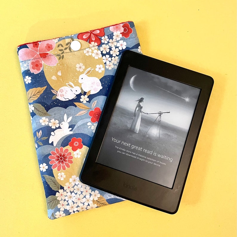 May include: A black Kindle e-reader with a screen displaying a woman and text that reads "Your next great read is waiting." Next to it is a blue fabric case with a floral and rabbit pattern.