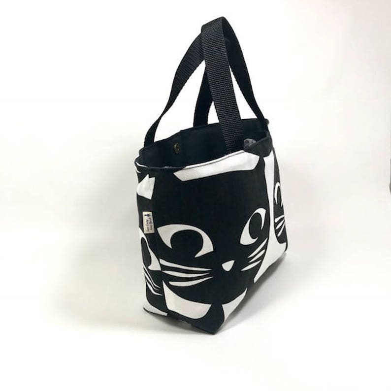 Puede incluir: Bolso tote negro y blanco con un dise&ntilde;o de cara de gato. El bolso tiene correas negras y un forro negro.