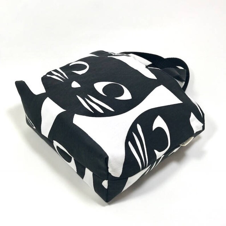 Puede incluir: Bolsa de tela negra y blanca con un patr&oacute;n repetido de gatos negros sobre fondo blanco. La bolsa tiene un asa negra.
