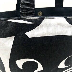 Puede incluir: Bolsa de tela negra y blanca con un dise&ntilde;o de gato. La bolsa tiene asas negras y una cara de gato blanca con ojos grandes.